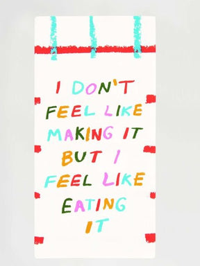 A white folded Blue Q tea towel with red and aqua grid lines, featuring colorful block letters reading "I DON'T FEEL LIKE MAKING IT BUT I FEEL LIKE EATING IT".