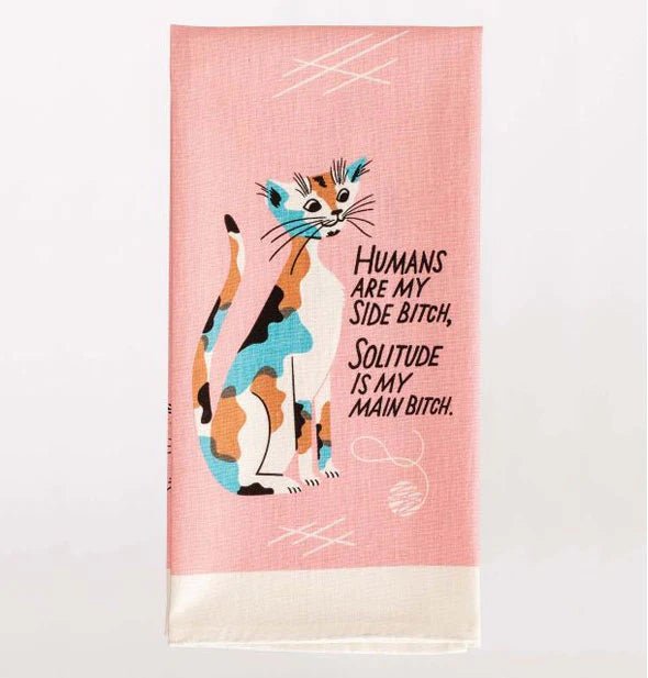 A pink folded Blue Q tea towel featuring a retro multi-colored cat illustration next to text reading "HUMANS ARE MY SIDE BITCH, SOLITUDE IS MY MAIN BITCH".
