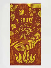 A rust-red folded Blue Q tea towel with yellow illustrations of a skillet, flames, vegetables, and text reading "I SAUTÉ. I'm fancy."