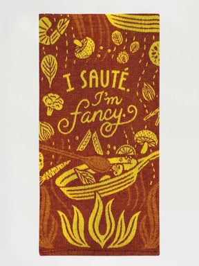 A rust-red folded Blue Q tea towel with yellow illustrations of a skillet, flames, vegetables, and text reading "I SAUTÉ. I'm fancy."