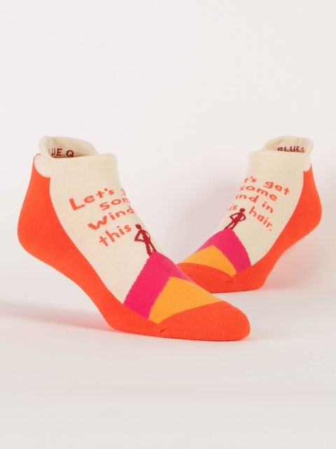 Pair of Blue Q low-cut sneaker socks in cream, orange, pink, and yellow featuring a hill illustration and text reading "Let's get some wind in this hair."