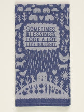 A folded blue and white woven Blue Q tea towel with folk-art illustrations of farm animals, rain, and text reading "SOMETIMES BLESSINGS LOOK A LOT LIKE BULLSHIT."
