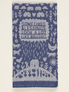A folded blue and white woven Blue Q tea towel with folk-art illustrations of farm animals, rain, and text reading "SOMETIMES BLESSINGS LOOK A LOT LIKE BULLSHIT."