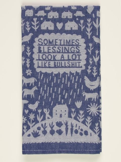 A folded blue and white woven Blue Q tea towel with folk-art illustrations of farm animals, rain, and text reading "SOMETIMES BLESSINGS LOOK A LOT LIKE BULLSHIT."