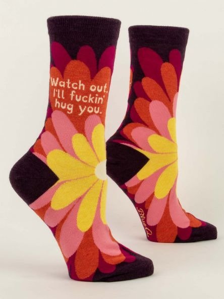 Short & Descriptive: Pair of Blue Q women's purple crew socks featuring a large retro flower in yellow, orange, and pink with text reading "Watch out. I'll fuckin' hug you."