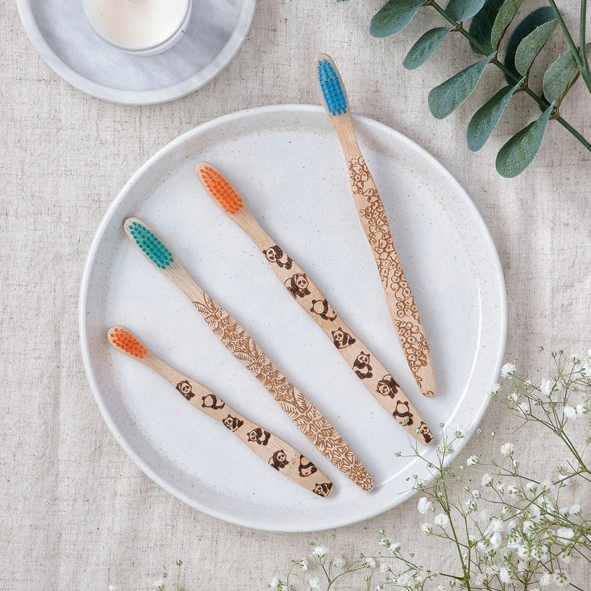 Brush It On Lassie kids bamboo toothbrushes with various animal handle engravings and multi-colored bristles displayed on a white ceramic plate with a textured tablecloth and green foliage