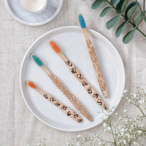 Brush It On Lassie kids bamboo toothbrushes with various animal handle engravings and multi-colored bristles displayed on a white ceramic plate with a textured tablecloth and green foliage