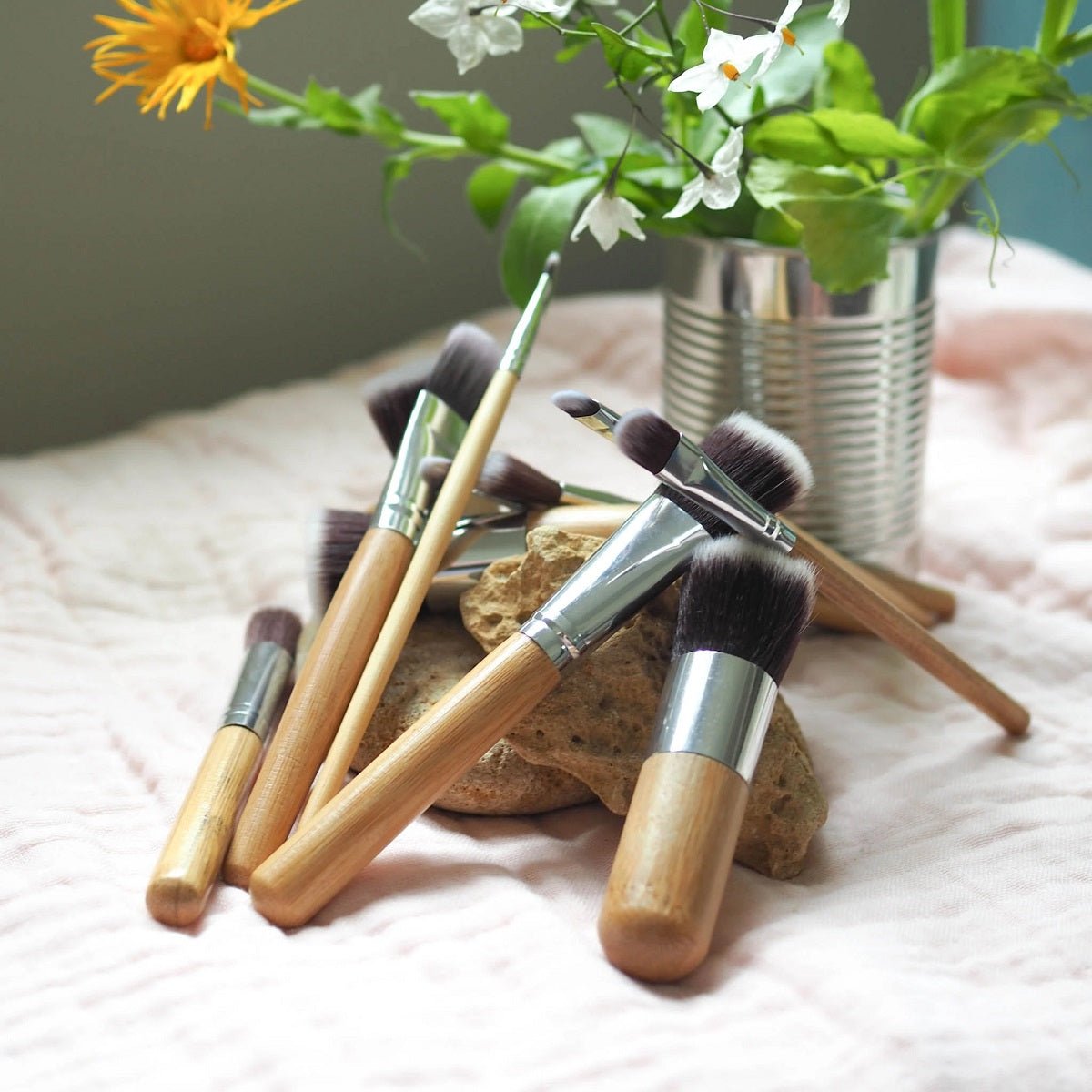 Brush It On 11-piece bamboo vegan makeup brush set arranged on a stone and pink fabric with a tin of fresh flowers in the background