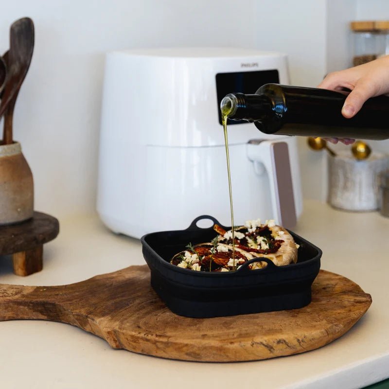 Olive oil being poured from a glass bottle onto stuffed mushrooms resting in a liquorice black Seed & Sprout silicone air fryer bowl.