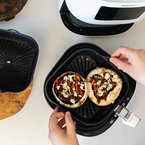 Hands lifting a liquorice black Seed & Sprout silicone air fryer bowl filled with stuffed mushrooms out of an air fryer basket.