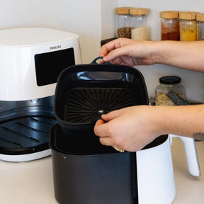 A pair of hands placing a liquorice black Seed & Sprout silicone air fryer bowl into the basket of a modern white air fryer on a kitchen counter.