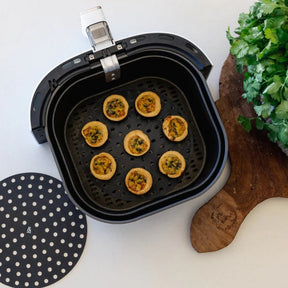 Mini quiches baking on a black perforated Seed & Sprout silicone air fryer mat inside an air fryer basket next to a second mat and a wooden cutting board with fresh herbs.