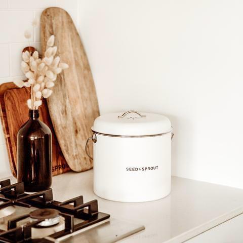 Seed & Sprout off-white mushroom compost bin placed on a kitchen countertop alongside wooden cutting boards and a dark amber glass bottle with dried lagurus grass.