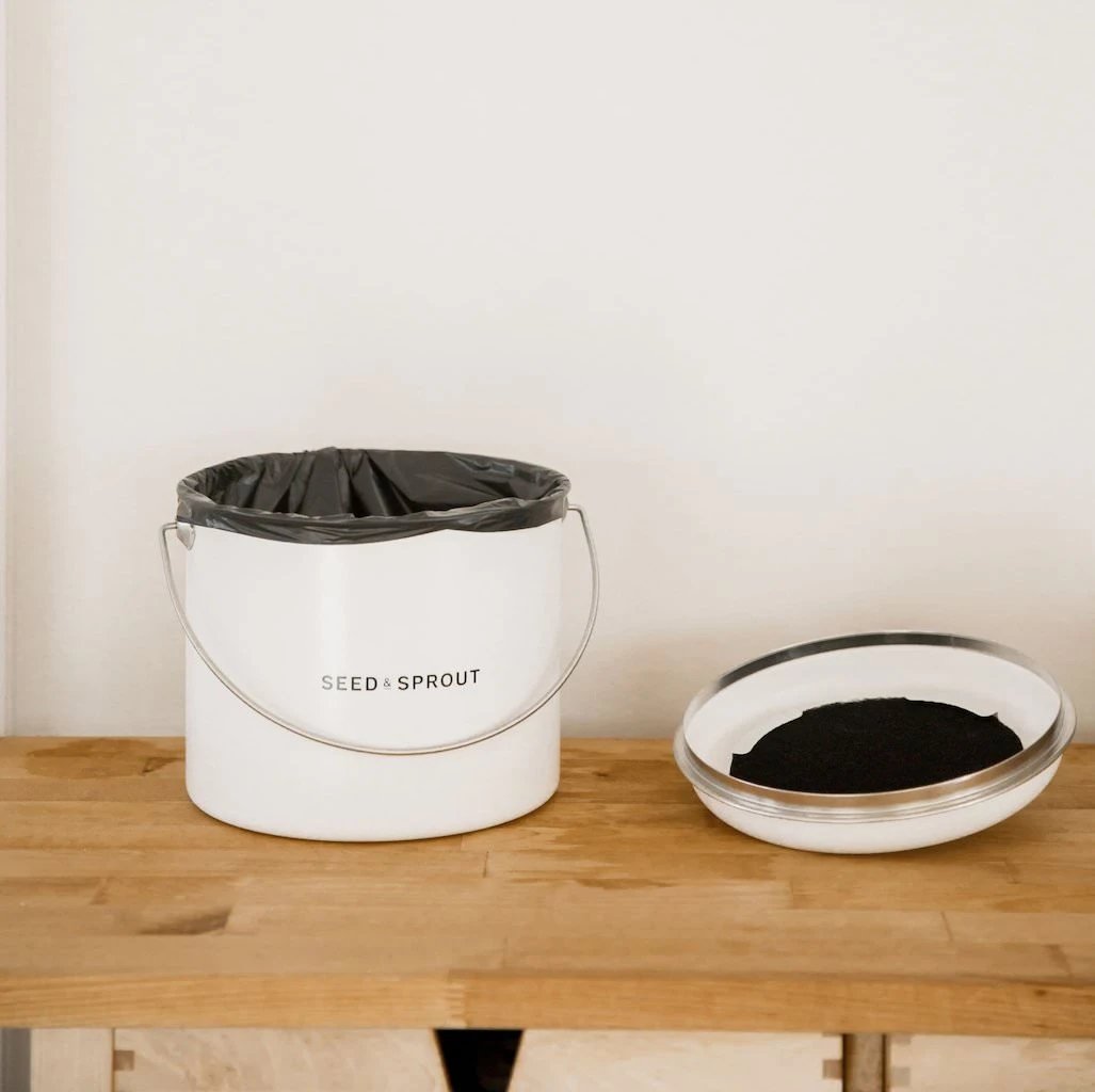 Seed & Sprout mushroom kitchen compost bin lined with a dark compostable bag, resting on a wooden tabletop next to its inverted lid displaying the charcoal filter.