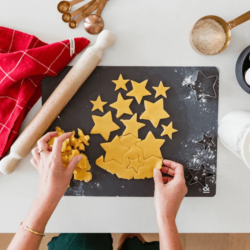 Flat lay view of star-shaped cookie dough cutouts on a dark graphite Seed & Sprout silicone baking mat alongside kitchen baking tools.