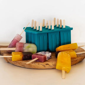 A collection of colorful homemade fruit popsicles resting on a wooden board next to the Seed & Sprout juniper silicone icy pole mould.