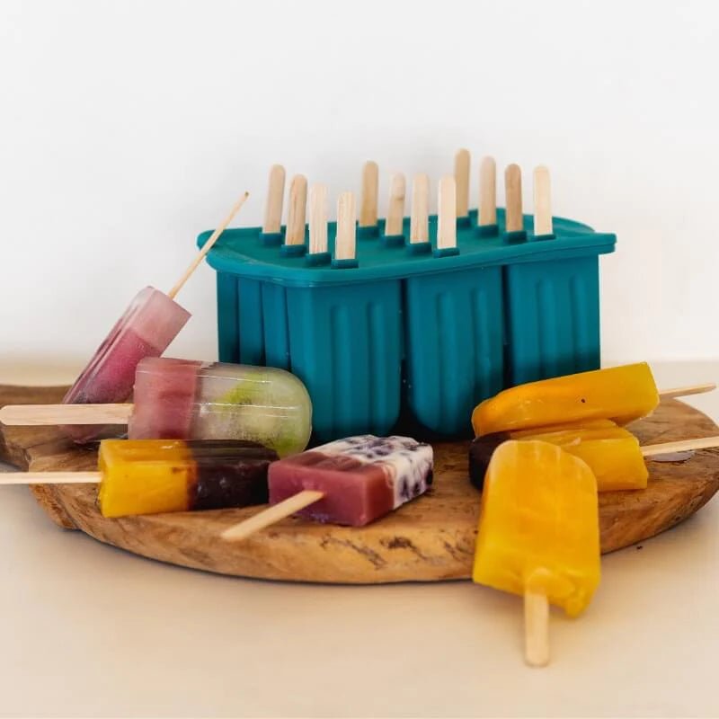 A collection of colorful homemade fruit popsicles resting on a wooden board next to the Seed & Sprout juniper silicone icy pole mould.