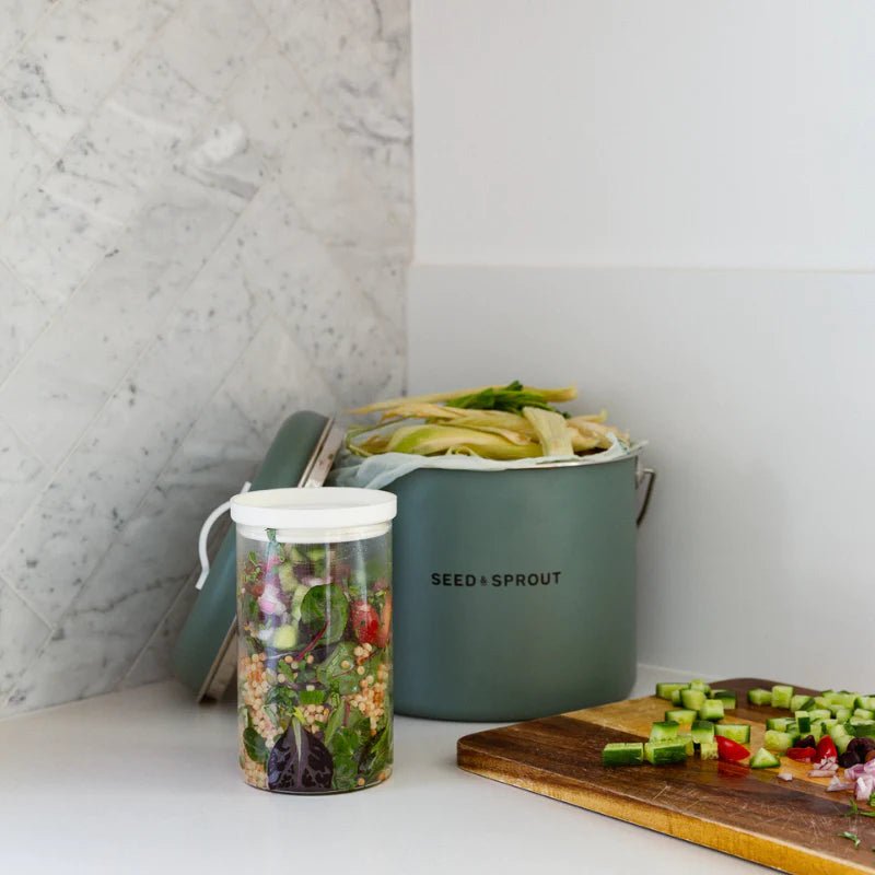 Seed & Sprout eucalyptus green kitchen compost bin filled with vegetable scraps alongside a meal-prep glass jar and a cutting board on a white countertop.