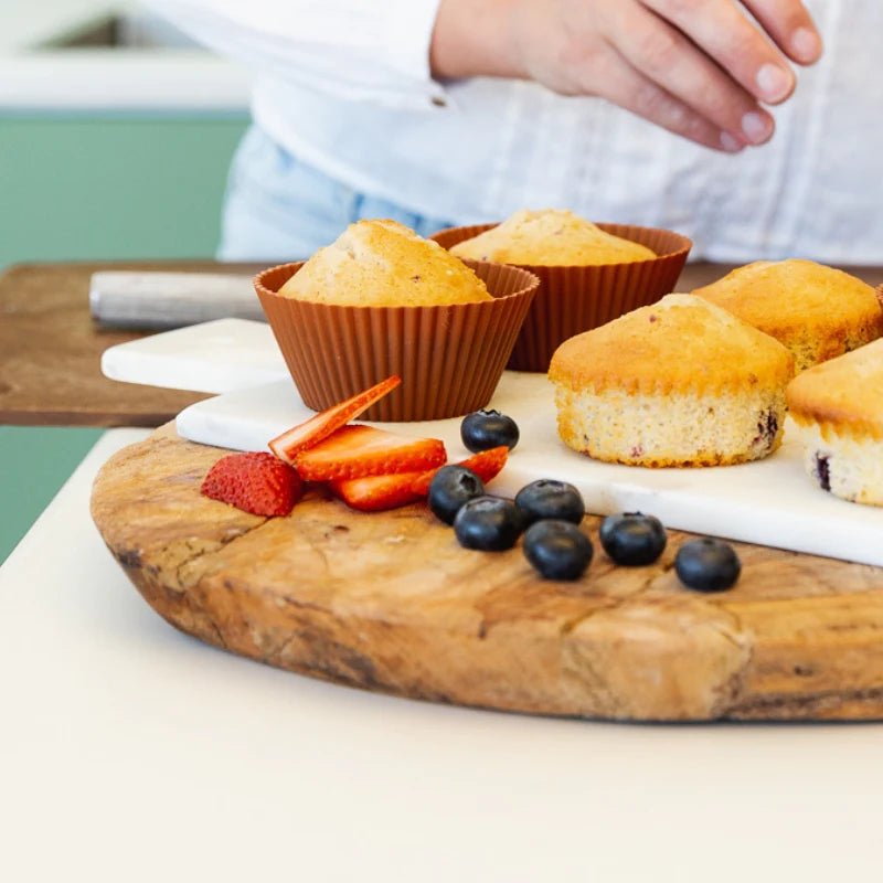Freshly baked muffins in Seed & Sprout cocoa silicone baking cups alongside unlined muffins, strawberries, and blueberries on a wooden cutting board.