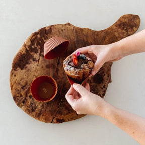 Seed & Sprout cocoa silicone baking cup being peeled away from a freshly baked gourmet muffin topped with berries on a rustic wooden cutting board.