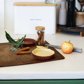 Seed & Sprout terracotta silicone mini pie dish filled with pastry dough sitting on a wooden cutting board alongside an apple, cinnamon sticks, and a white bread bin.