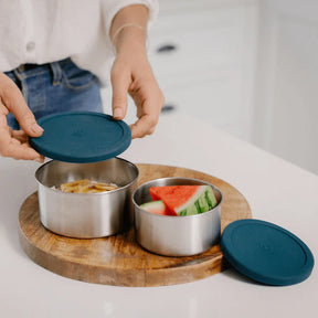 Seed & Sprout stainless steel nesting containers in forest green being opened in a kitchen, filled with sliced fruit on a wooden board.