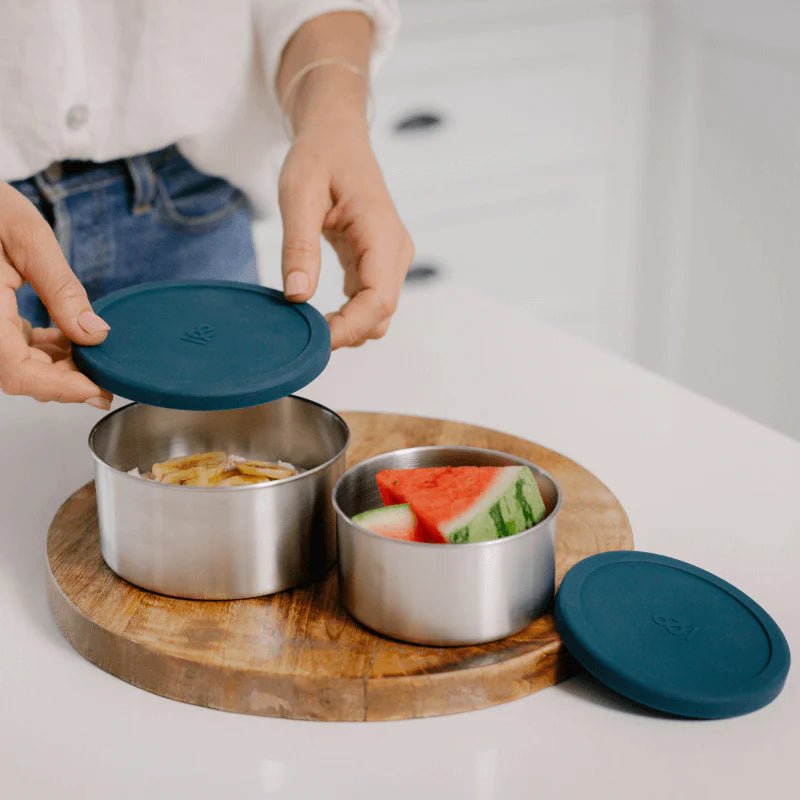 Seed & Sprout stainless steel nesting containers in forest green being opened in a kitchen, filled with sliced fruit on a wooden board.
