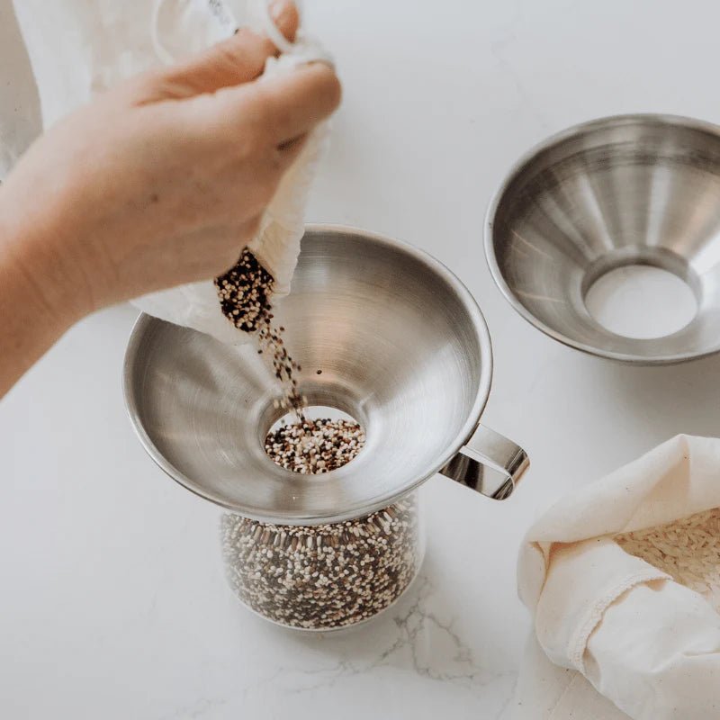 Seed & Sprout stainless steel funnel being used to pour grains from a reusable cotton produce bag into a glass jar on a white kitchen counter.